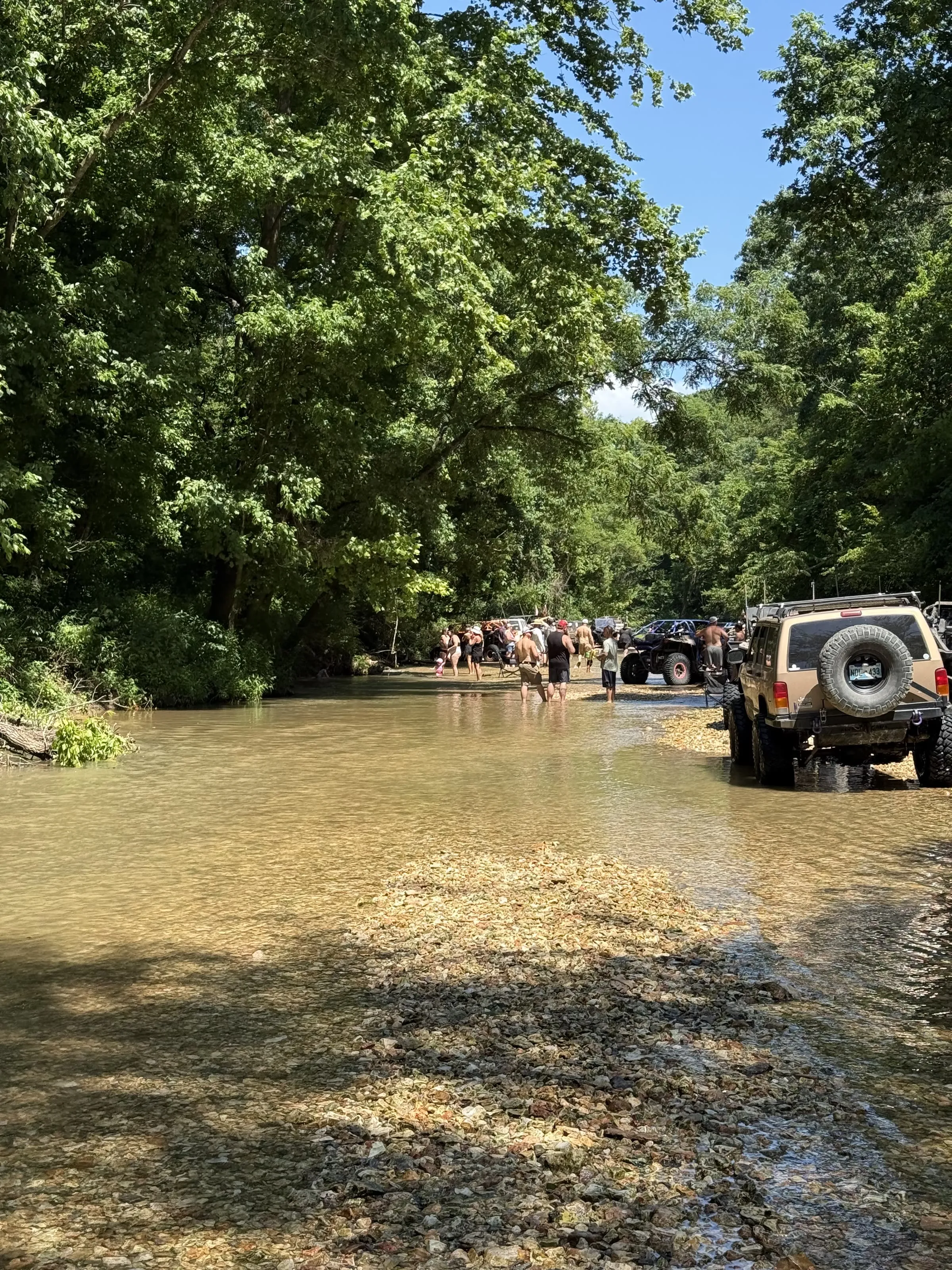 A group gathered at a shady creek crossing with a Jeep parked at the water's edge