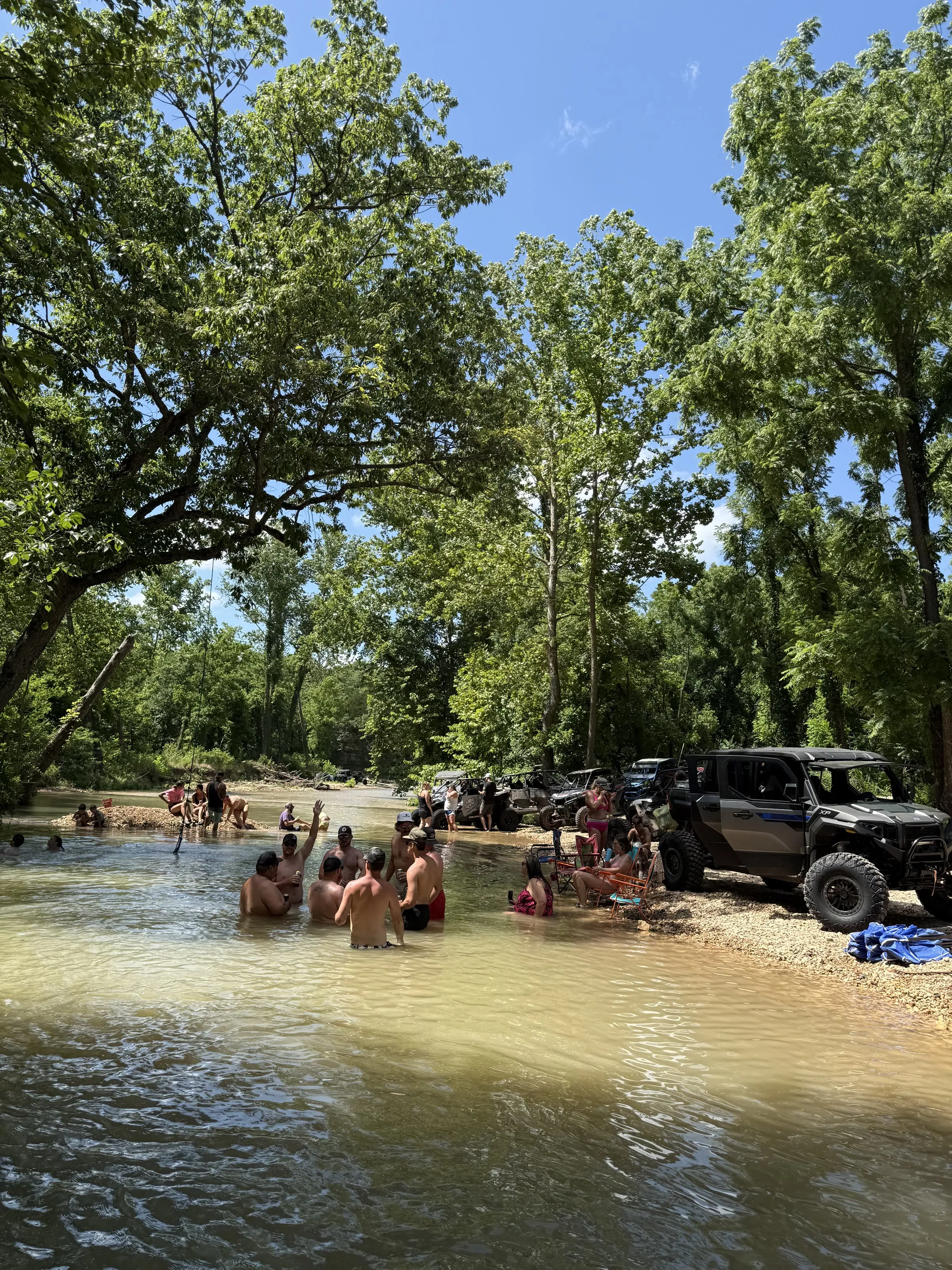A group swimming and wading at a Shultz Creek swim hole with a rope swing overhead and UTVs parked at the water's edge