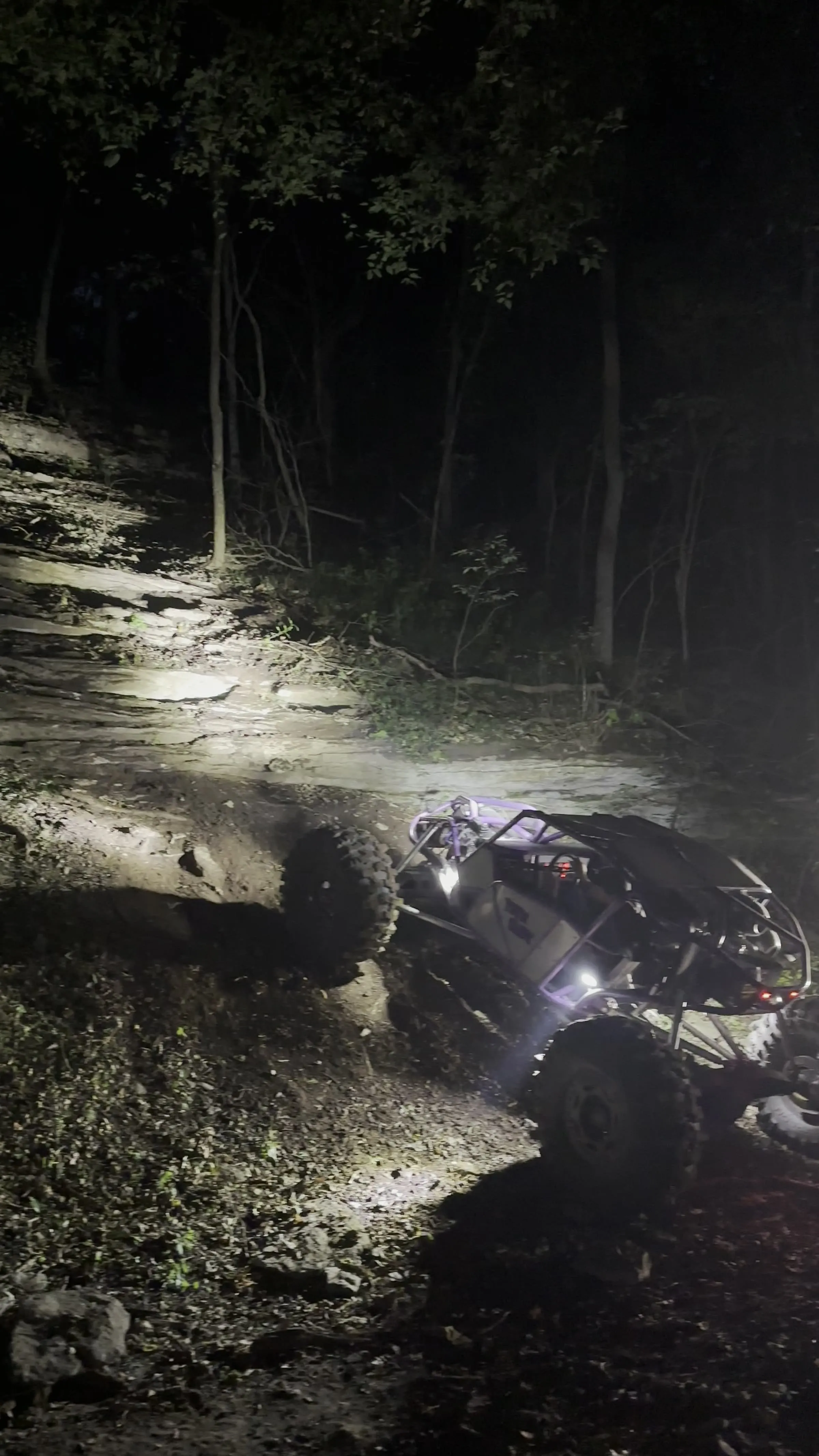A rock crawler buggy climbing a rocky ledge at night, headlights cutting through the dark
