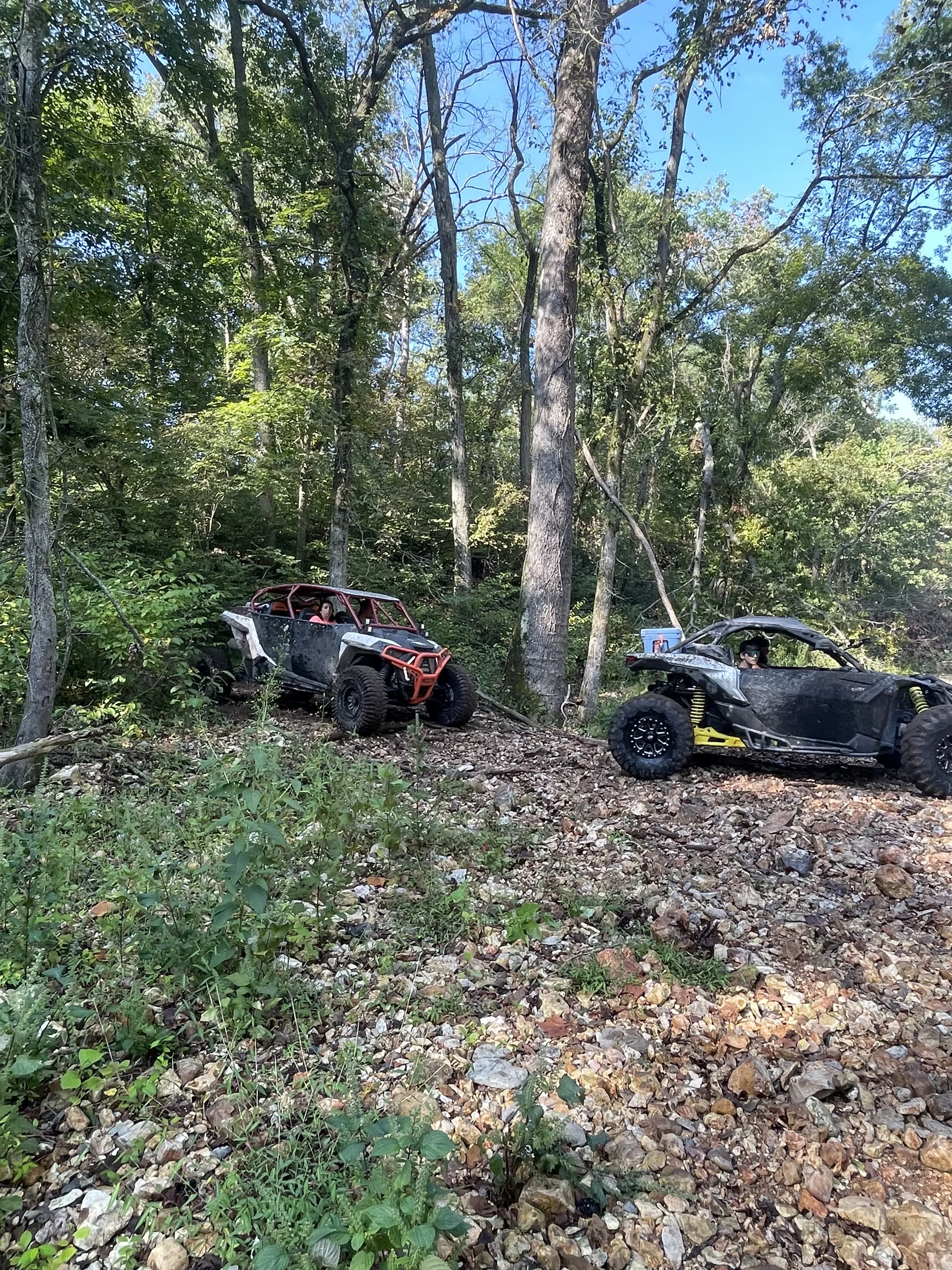 Two mud-covered UTVs parked on a rocky wooded hillside trail