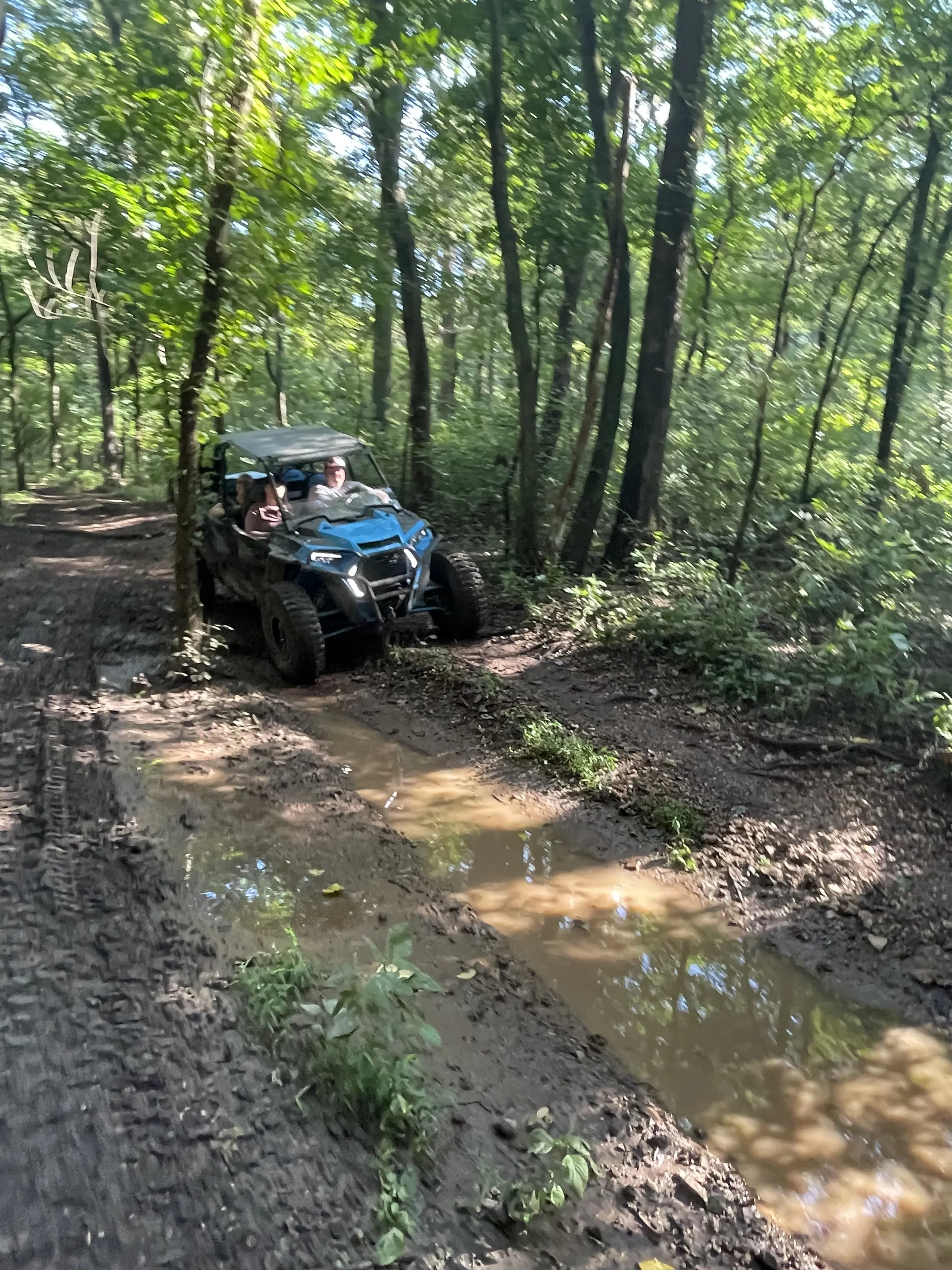 A UTV with two riders crossing a muddy trail puddle